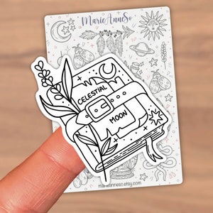 May include: Black and white line art sticker of a closed book with a celestial moon design. The book has a strap with the words "Celestial Moon" written on it. A sprig of flowers is on the left side of the book.