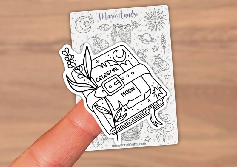 May include: Black and white line art sticker of a closed book with a celestial moon design. The book has a strap with the words "Celestial Moon" written on it. A sprig of flowers is on the left side of the book.