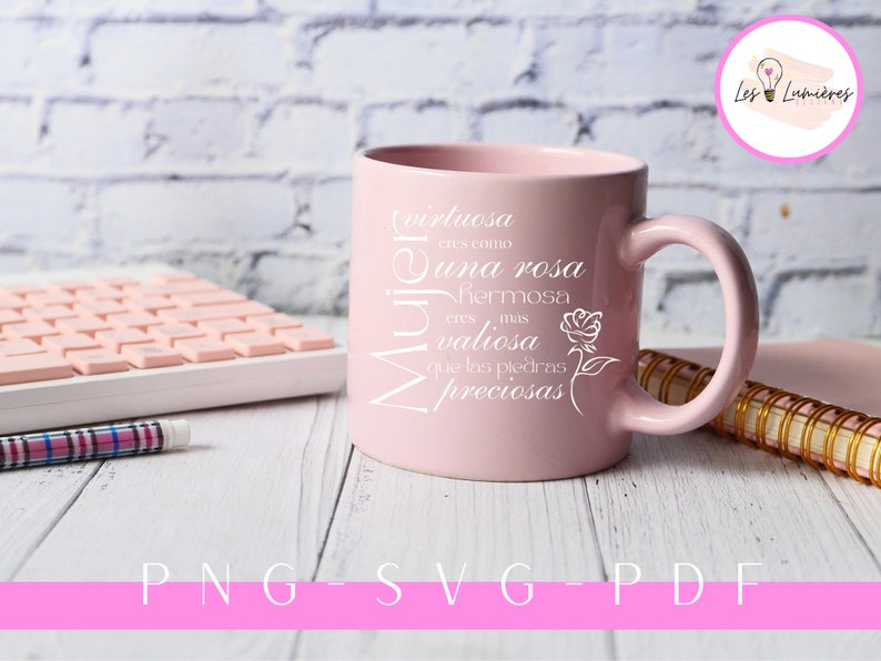 Op de afbeelding: Een roze keramische mok met een wit ontwerp met de tekst "Virtuosa eres como una rosa hermosa eres mas valiosa que las piedras preciosas" en een kleine roos afbeelding. De mok staat op een wit houten oppervlak met een toetsenbord en een pen.