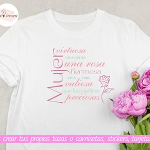 Op de afbeelding: Een wit T-shirt met een roze en groen ontwerp met de tekst "virtuosa eres como una rosa hermosa eres mas valiosa que las piedras preciosas" in het Spaans. De tekst is verticaal gerangschikt en het woord "Mujer" staat ondersteboven. Het T-shirt ligt op een lichtgrijze ondergrond met een boeket roze pioenen in de hoek.