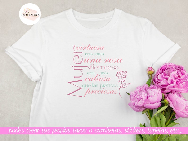 Op de afbeelding: Een wit T-shirt met een roze en groen ontwerp met de tekst "virtuosa eres como una rosa hermosa eres mas valiosa que las piedras preciosas" in het Spaans. De tekst is verticaal gerangschikt en het woord "Mujer" staat ondersteboven. Het T-shirt ligt op een lichtgrijze ondergrond met een boeket roze pioenen in de hoek.