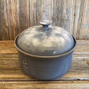 Pode incluir: Uma cloche de pão de cerâmica cinza com tampa. A tampa tem um esmalte branco salpicado e uma pega no topo. A cloche tem três furos no lado.