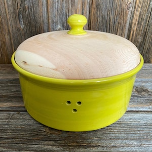 Könnte beinhalten: Ein runder, limettengrüner Keramik-Brotbacktopf mit einem hellen Holzdeckel und einem passenden gelben Knauf. Der Topf hat drei kleine Löcher an der Seite. Der Deckel ist aus hellem Holz.