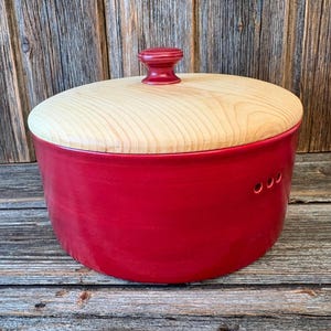 Può includere: Un portapane in ceramica rossa con coperchio e pomello in legno chiaro. Il portapane ha tre piccoli fori sul lato. Il coperchio è di forma ovale e si adatta perfettamente sopra il contenitore rosso. Il portapane è su una superficie di legno.