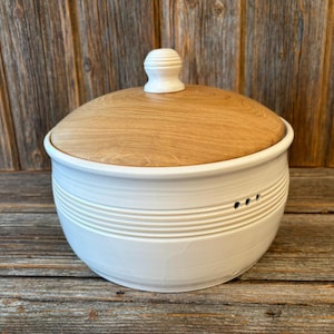 Puede incluir: Un cuenco de pan de cerámica blanca con tapa de madera. El cuenco tiene estrías horizontales y tres pequeños agujeros. La tapa es de color marrón claro con un pomo blanco. El cuenco está sobre una superficie de madera.