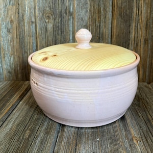 Pode incluir: Uma tigela de cerâmica rosa claro com tampa de madeira. A tampa é amarelo claro com um pequeno botão redondo rosa claro. A tigela tem pequenos orifícios perto da parte superior e está sobre uma superfície de madeira.
