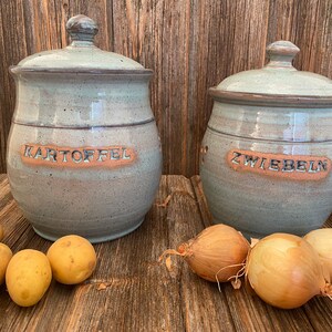 Peut inclure: Deux pots de stockage en céramique avec couvercles, l'un étiqueté "KARTOFFEL" et l'autre "ZWIEBELN". Le pot étiqueté "KARTOFFEL" a des pommes de terre devant lui, et le pot étiqueté "ZWIEBELN" a des oignons devant lui.