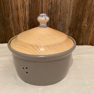 Può includere: Contenitore per pane in ceramica grigia con coperchio in legno e pomo in ceramica bianca. Il coperchio ha un motivo floreale blu e bianco.