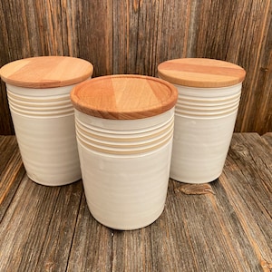 Pode incluir: Três potes de cerâmica branca com tampas de madeira. Os potes têm um design sutil em listras e são perfeitos para guardar mantimentos de cozinha.