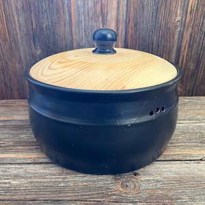 Könnte beinhalten: Eine schwarze Brotdose aus Keramik mit einem Holzdeckel. Der Deckel hat einen kleinen Knopf oben.