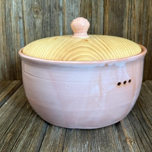 Könnte beinhalten: Eine handgefertigte Keramikschale mit hellrosa Glasur und einem Holzdeckel. Der Deckel ist hell gefärbt und hat ein kreisförmiges Maserungsmuster. Die Schale hat drei kleine Löcher an der Seite und einen kleinen Knopf auf dem Deckel.