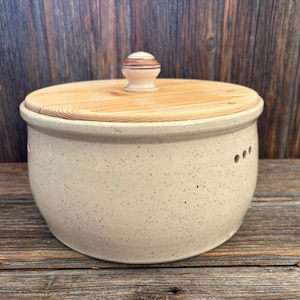 Pode incluir: Um pote de pão de cerâmica creme salpicado com uma tampa de madeira clara e um pequeno botão decorativo. O pote tem três pequenos orifícios na lateral. A tampa é feita de madeira clara e está sobre uma superfície de madeira.