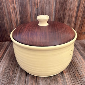 Op de afbeelding: Een gele keramische container met een houten deksel. Het deksel heeft een kleine knop bovenop. De container heeft een getextureerd oppervlak en staat op een houten oppervlak.