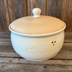 Può includere: Un contenitore per l'aglio in ceramica con una glassa giallo chiaro e un coperchio in legno. Il contenitore ha tre piccoli fori e un disegno intagliato sul lato. Il coperchio ha un piccolo pomello rotondo. Il contenitore è su una superficie di legno.