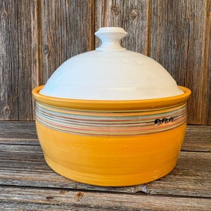 Può includere: Una ciotola da portata in ceramica con coperchio. La ciotola è di un giallo brillante con una fascia di strisce orizzontali colorate al centro. Il coperchio è bianco con una piccola maniglia sulla parte superiore. La ciotola è su una superficie di legno.