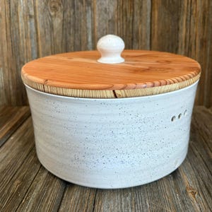 May include: A round, speckled white ceramic container with a wooden lid. The lid is made of light-coloured wood and has a small, white knob. The container has three small holes on the side.