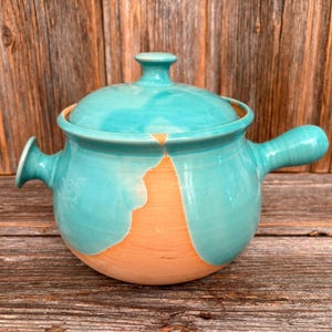 Op de afbeelding: Een turquoise keramische ovenschaal met een deksel en twee handvatten. Het onderste deel van de schaal heeft een natuurlijke kleikleur, waardoor een tweekleurig effect ontstaat. De schaal staat op een houten oppervlak.