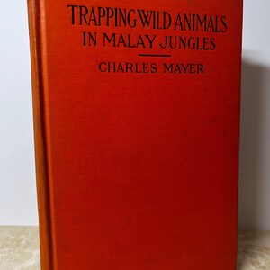 May include: A red hardback book with the title "Trapping Wild Animals in Malay Jungles" and the author's name "Charles Mayer" printed in black.