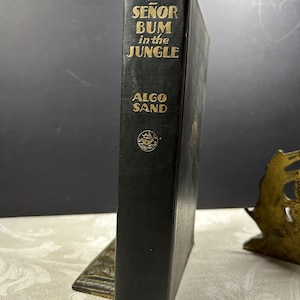 May include: A vintage hardcover book titled "Señor Bum in the Jungle" by Algo Sand. The book has a black cover with gold lettering and a decorative emblem. The book is standing upright, showcasing its spine.