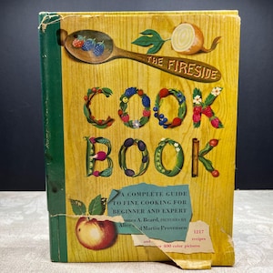 May include: Vintage cookbook titled "The Fireside Cook Book" with a yellow cover. The title is spelled out with colorful illustrations of fruits and vegetables. The cover also features a wooden spoon with berries and an onion. The book is a complete guide to fine cooking.