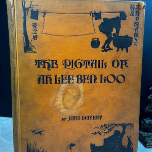 以下が含まれることがあります： 表紙に女性の黒いシルエットが描かれた茶色の本。本のタイトルは「The Pigtail of Ak Lee Ben Loo」で、ジョン・ベネット著です。
