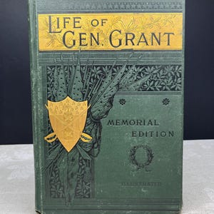 Puede incluir: Un libro antiguo titulado "Life of Gen. Grant" con una cubierta verde oscuro con letras y detalles dorados. La portada presenta un escudo dorado con espadas cruzadas y las palabras "Memorial Edition" e "Illustrated".