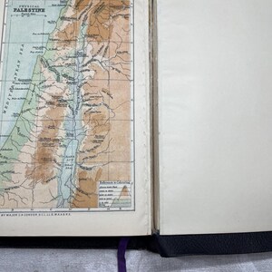 Puede incluir: Libro abierto con un mapa detallado de Palestina, con detalles topogr&aacute;ficos y el mar Mediterr&aacute;neo. El mapa es en color, con tonos marrones, verdes y azules. El libro tiene una cubierta negra y una cinta morada como marcador.