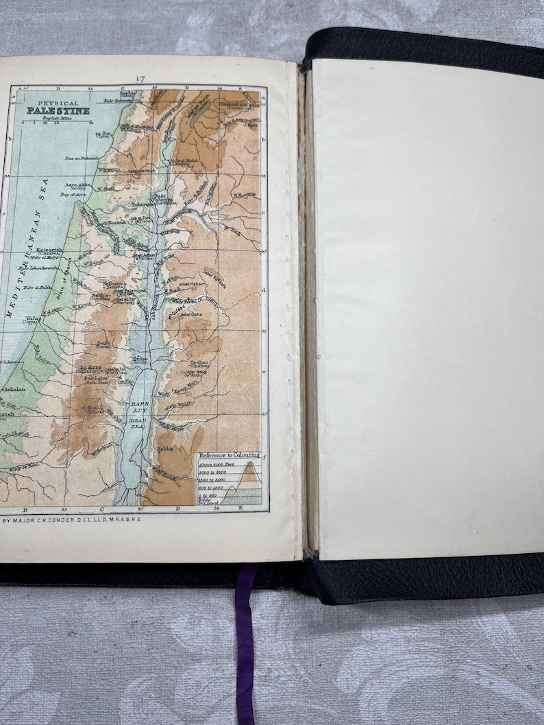 Puede incluir: Libro abierto con un mapa detallado de Palestina, con detalles topogr&aacute;ficos y el mar Mediterr&aacute;neo. El mapa es en color, con tonos marrones, verdes y azules. El libro tiene una cubierta negra y una cinta morada como marcador.