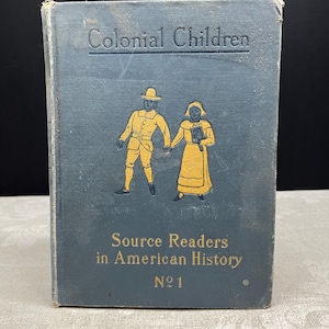 Pode incluir: Um livro vintage intitulado "Colonial Children" com uma capa azul. A capa apresenta ilustrações de um homem e uma mulher em trajes amarelos, de mãos dadas. O texto "Source Readers in American History No 1" também é visível.