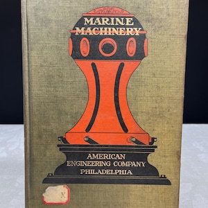 May include: A vintage book titled "MARINE MACHINERY" with an orange and black illustration. The book cover also features the text "AMERICAN ENGINEERING COMPANY PHILADELPHIA" and a small label with the letter "K". The book has a textured, olive green cover.