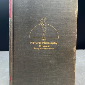May include: A vintage book titled "The Natural Philosophy of Love" by Remy de Gourmont. The cover is dark gray with gold lettering and a stylized illustration of a woman. The spine is red.