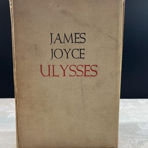 Puede incluir: Un libro de tapa dura titulado "Ulysses" de James Joyce. La portada es de color beige claro, con el nombre del autor y el título del libro impresos en negro y rojo. El libro parece ser antiguo, con algo de desgaste visible en la portada.