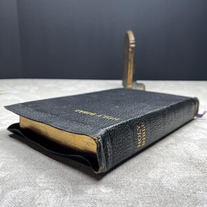 Puede incluir: Una Santa Biblia encuadernada en cuero negro con bordes dorados y letras doradas. El libro est&aacute; abierto sobre una superficie estampada, con un peque&ntilde;o objeto decorativo dorado en el fondo. Una cinta morada de marcador es visible.