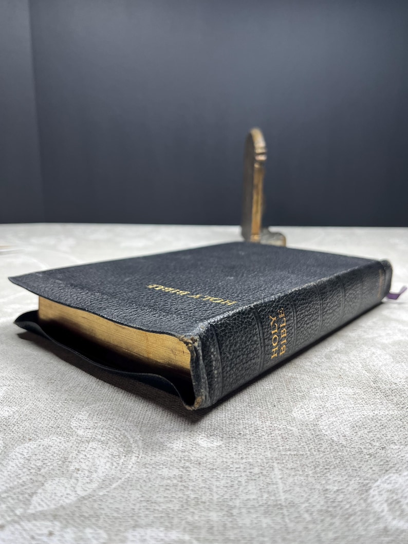 Puede incluir: Una Santa Biblia encuadernada en cuero negro con bordes dorados y letras doradas. El libro est&aacute; abierto sobre una superficie estampada, con un peque&ntilde;o objeto decorativo dorado en el fondo. Una cinta morada de marcador es visible.