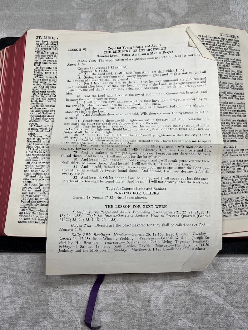 Puede incluir: Una Biblia abierta con una cinta morada como marcador, que muestra texto del Libro de Lucas y una lecci&oacute;n titulada "El Ministerio de la Intercesi&oacute;n". Las p&aacute;ginas est&aacute;n envejecidas, con texto y un plan de lecci&oacute;n visibles.