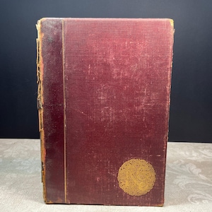 Könnte beinhalten: Ein altes gebundenes Buch mit einem dunkelroten Stoffeinband. Der Buchrücken besteht aus einem dunkleren, abgenutzten lederartigen Material mit sichtbaren Abnutzungserscheinungen. Auf dem Vorderdeckel befindet sich ein goldenes Emblem.
