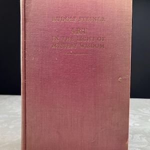 Może przedstawiać: Stara książka z wyblakłą czerwoną okładką. Tytuł "ART IN THE LIGHT OF MYSTERY WISDOM" jest wytłoczony złotymi literami. Imię i nazwisko autora, RUDOLF STEINER, również jest złote. Grzbiet książki jest widoczny, a krawędzie noszą ślady zużycia.