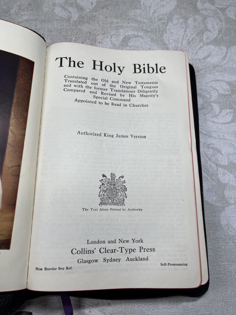 Puede incluir: Libro abierto titulado "The Holy Bible" con texto negro. La p&aacute;gina del t&iacute;tulo incluye el texto "Authorized King James Version" y la informaci&oacute;n del editor. El libro tiene un borde rojo y est&aacute; encuadernado en negro.