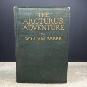 Puede incluir: Un libro de tapa dura titulado "The Arcturus Adventure" de William Beebe. El libro tiene una cubierta verde oscuro con letras doradas. El libro está cerrado y de pie.