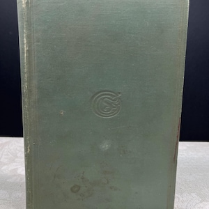 May include: A vintage book with a faded green cover. The cover has a simple embossed design in the center and a rectangular border. The edges of the book show signs of wear, suggesting its age. The book is standing upright.
