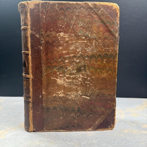 May include: An antique book with a worn, marbled cover in shades of brown, red, and green. The spine is a darker brown leather, and the edges show signs of age. The book's cover has a decorative pattern, and the overall aesthetic suggests a vintage item.