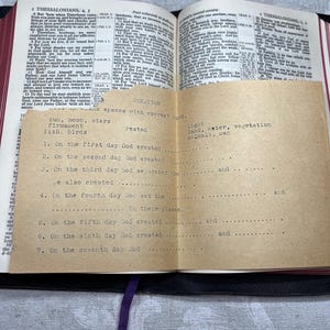 Puede incluir: Una Biblia abierta con un inserto de papel beige que detalla la historia de la creaci&oacute;n. El inserto tiene texto mecanografiado con espacios en blanco para completar. Una cinta morada marca una p&aacute;gina. Las p&aacute;ginas de la Biblia est&aacute;n amarillentas.