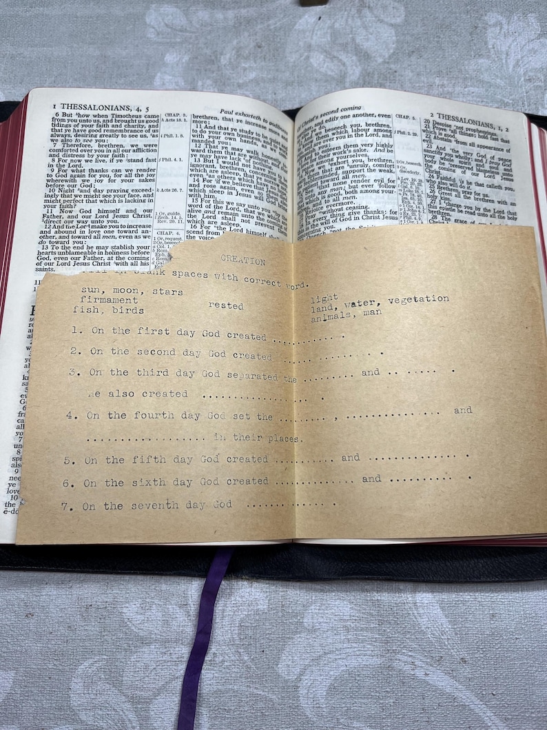 Puede incluir: Una Biblia abierta con un inserto de papel beige que detalla la historia de la creaci&oacute;n. El inserto tiene texto mecanografiado con espacios en blanco para completar. Una cinta morada marca una p&aacute;gina. Las p&aacute;ginas de la Biblia est&aacute;n amarillentas.