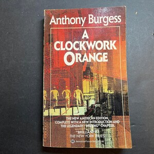 May include: A paperback book titled A Clockwork Orange by Anthony Burgess. The cover features a red and orange abstract background with silhouettes of four figures walking. Text includes The New American Edition and a New York Times quote.