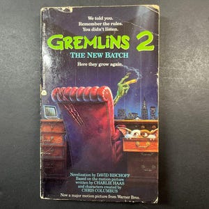 Pode incluir: Um livro de bolso intitulado Gremlins 2 The New Batch de David Bischoff. A capa apresenta uma mão verde com garras segurando um charuto sobre uma cadeira de escritório de couro vermelho, com uma pequena criatura espreitando de uma gaveta.