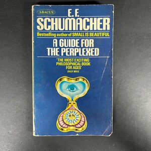May include: A paperback book titled A Guide for the Perplexed by E.F. Schumacher with a dark blue cover. The cover features the Abacus logo, the text Bestselling author of SMALL IS BEAUTIFUL, and a gold-toned graphic of an eye and a compass.