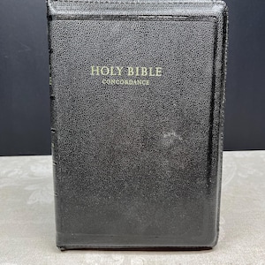 May include: A black leather-bound Holy Bible with gold lettering that reads "Holy Bible Concordance." The book has a textured surface and is standing upright. The background is a dark color.