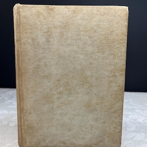 May include: A vintage book with a beige linen cover. The cover shows signs of age with discoloration and wear. The book is upright, with the spine and front cover visible, set against a dark background.