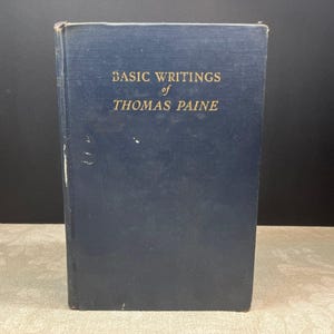 Op de afbeelding: Een vintage hardcover boek met de titel "Basic Writings of Thomas Paine". Het boek heeft een donkerblauwe kaft met gouden letters. De rug vertoont slijtage, wat duidt op de leeftijd. Het boek staat rechtop en toont de titel.