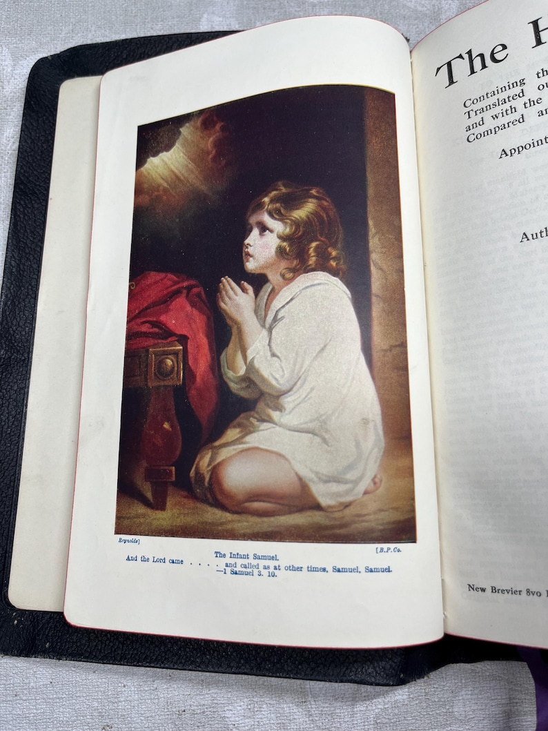 Puede incluir: Libro abierto con una ilustraci&oacute;n en color de un ni&ntilde;o arrodillado en oraci&oacute;n. El ni&ntilde;o lleva una prenda blanca. El texto debajo de la imagen dice "The Infant Samuel" y "And the Lord came...". El libro est&aacute; encuadernado en cuero negro.